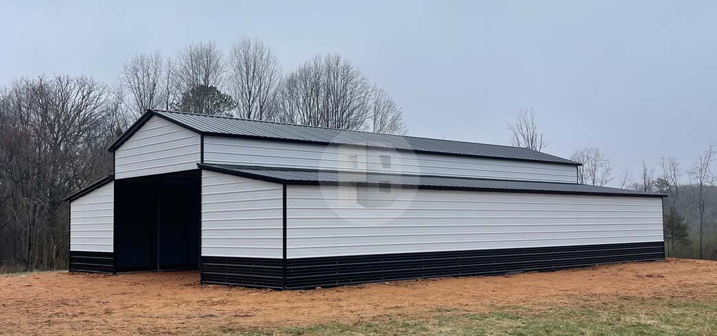 Long white metal barn with a black roof and black wainscot, one open bay on the near end, standing on bare red dirt