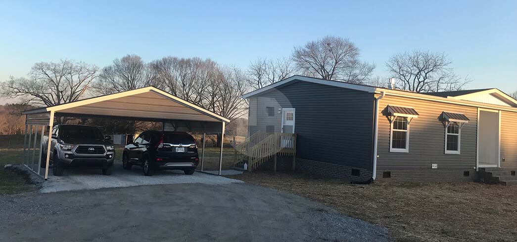 Open-sided metal carport with a tan gable roof sheltering two vehicles on a gravel drive beside a house